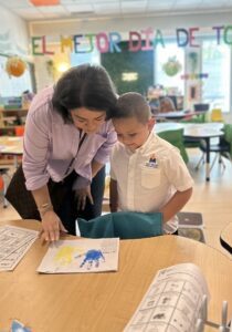 Teacher guides a young student as they review artwork at a classroom table during an ECC Open House.